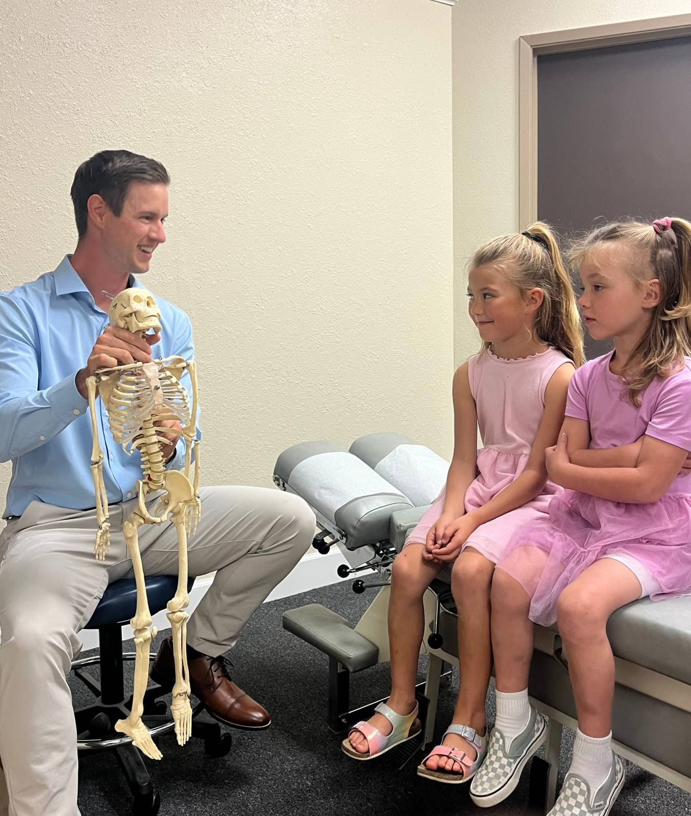 Dr. Hill, smiling, showing a skeleton model to two young girls sitting on an adjusting table